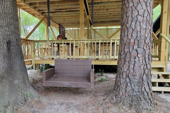 Wooden treehouse platform with a swing bench below and tall trees around.