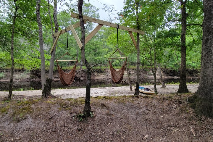 Two hanging chairs in a wooded area beside a creek.