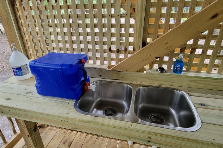 Outdoor sink setup with two basins, a blue water container, and cleaning supplies on a wooden counter.