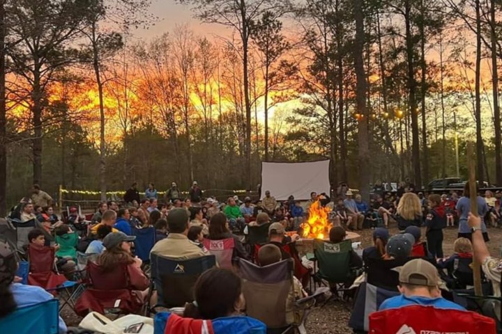 People seated around a campfire during sunset in a forest, with a screen set up in the background.
