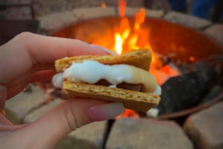 Person holding a s'more with marshmallow by a campfire.